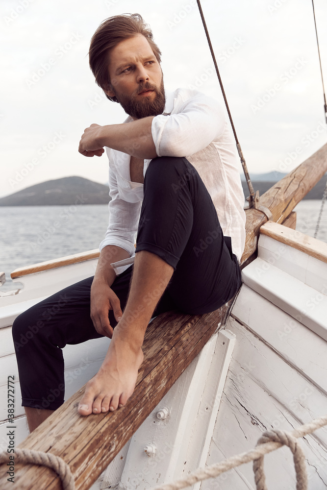 handsome man with a beard at sunset on a yacht wearing a white linen shirt and pants with a brutal expression