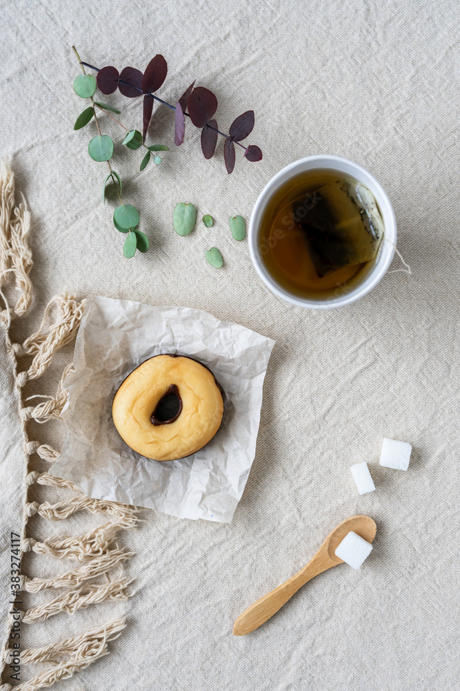 Ein Pappbecher mit Kräutertee und ein Donat auf einem Backpapier. Leinen Tischtuch, Flat lay, Würfelzucker, Teepause. Ein Pappbecher mit Kräutertee und ein Donat auf einem Backpapier. Leinen Tischtuch, Flat lay, Würfelzucker, Teepause.