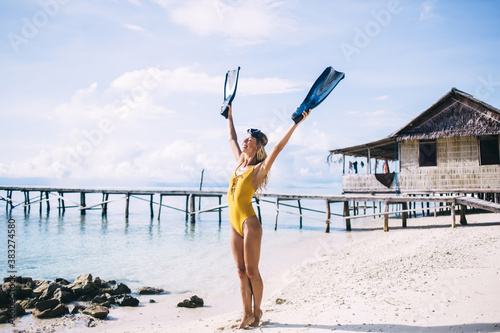Joyful woman raising swim flippers while satisfy at sea coastline of tropical island on Bahamas, happy female swimmer rejoicing during leisure time for practicing snorkeling hobby on Raja Ampat