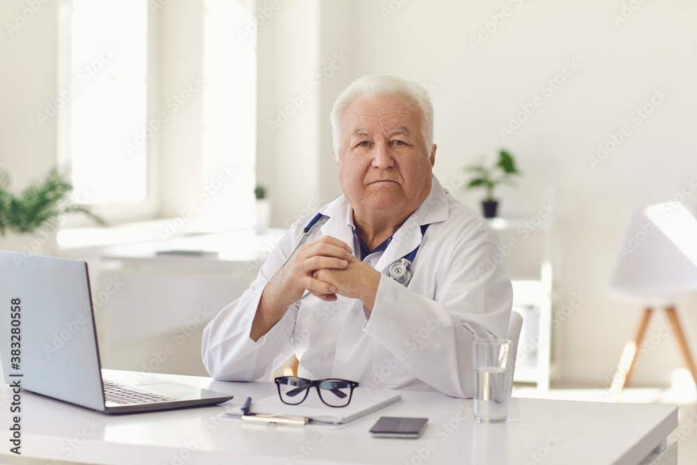 Serious senior physician sitting at desk with laptop in hospital office looking at camera