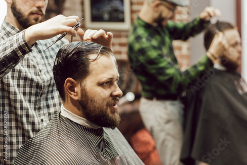 Haircut in the barbershop. Close up view of young bearded man getting haircut while sitting in chair at barbershop. Hairdresser cuts hair with scissors and a comb