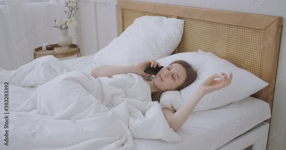 Young caucasian woman sitting on the bed in the morning feels bad. A young woman is making a smartphone call to her doctor. Young caucasian woman sitting on the bed in the morning feels bad. A young woman is making a smartphone call to her doctor.