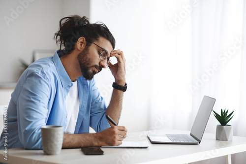 Concentrated Indian Freelancer Guy Noting Ideas To Notepad, Working At Home Office