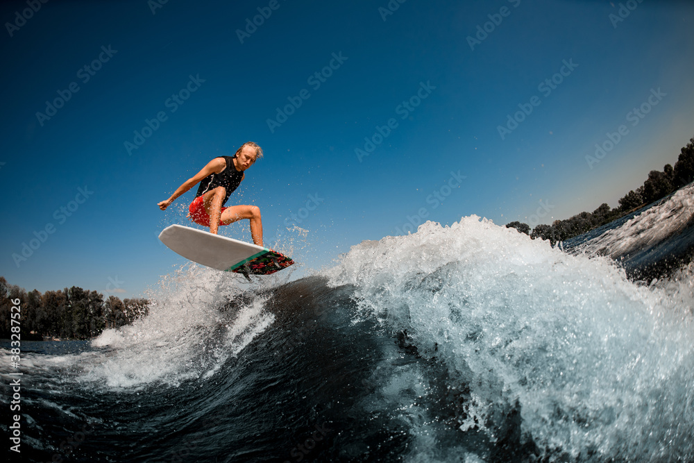 man bounces on surf style wakeboard over splashing wave against blue sky