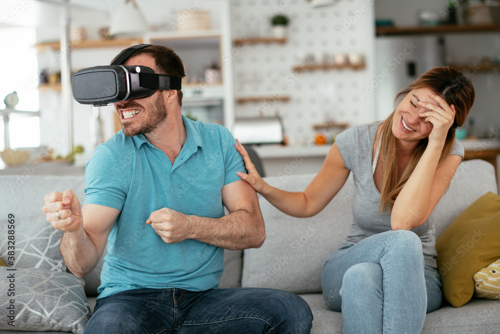 Fototapeta premium Smiling young man using VR headset at home on couch. Woman and her husband enjoying virtual reality in apartment..