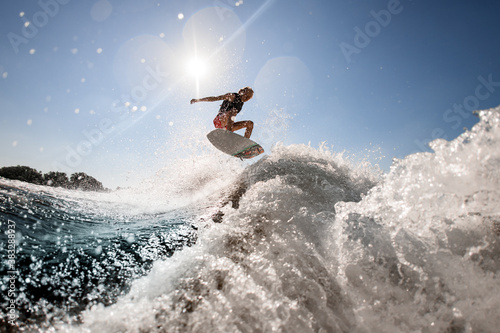 man professionally rides and bounces on surf style wakeboard over splashing wave against blue sky
