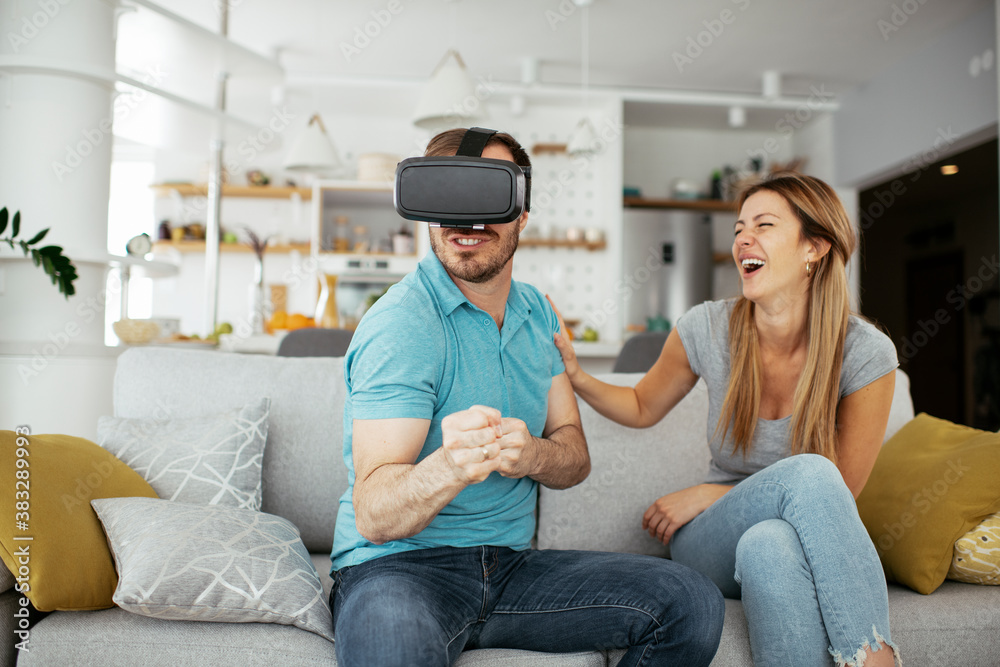 Smiling young man using VR headset at home on couch. Woman and her husband enjoying virtual reality in apartment.. Smiling young man using VR headset at home on couch. Woman and her husband enjoying virtual reality in apartment..