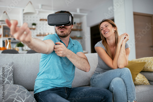Smiling young man using VR headset at home on couch. Woman and her husband enjoying virtual reality in apartment..