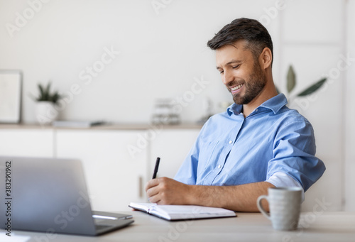 Smiling Handsome Businessman Taking Notes To Notepad At Workplace In White Office