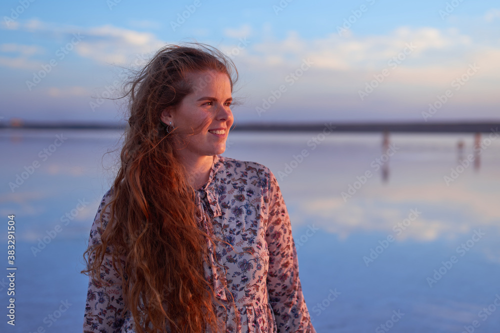 Beautiful smiling young woman with flying red hair on the ocean coast at sunset time