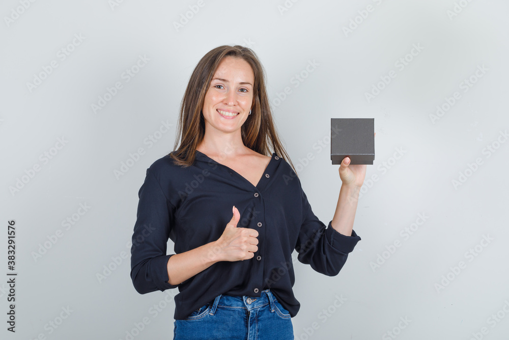 Naklejka premium Young woman in black shirt, jeans shorts holding watch box with thumb up and looking glad , front view.