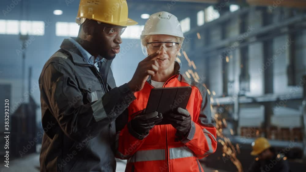 Two Heavy Industry Engineers Stand in Steel Metal Manufacturing Factory, Use Digital Tablet Computer and Have a Discussion. Black African American Industrial Specialist Talk to Female Technician. Two Heavy Industry Engineers Stand in Steel Metal Manufacturing Factory, Use Digital Tablet Computer and Have a Discussion. Black African American Industrial Specialist Talk to Female Technician.