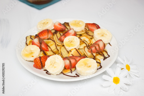 Trendy food - pancake cereal. Heap of mini cereal pancakes on a white plate on a white background. Strawberry slices on a plate, drizzled with syrup. Camomile flowers nearby.
