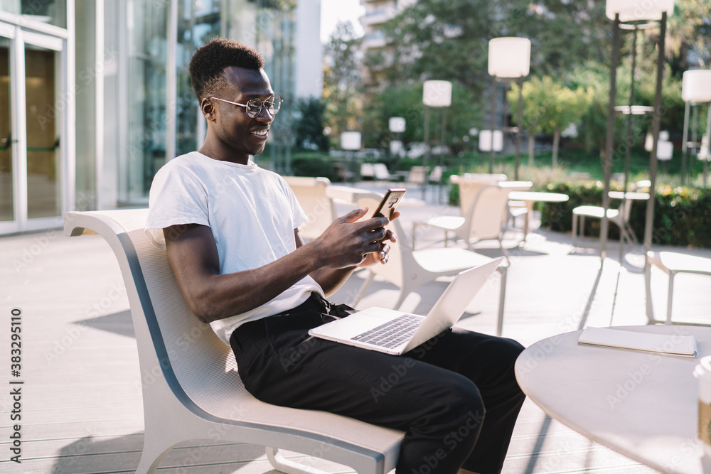 Smiling prosperous dark skinned male freelancer working via modern technology on cafe terrace on sunny day, happy african american 20s hipster guy reading messages on smartphone on remote job Smiling prosperous dark skinned male freelancer working via modern technology on cafe terrace on sunny day, happy african american 20s hipster guy reading messages on smartphone on remote job