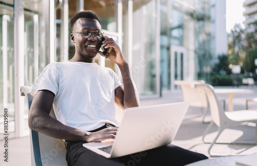 Smiling dark skinned hipster guy in eyewear for vision correction working remotely and talking on smartphone in roaming, positive african american male blogger on cafe terrace making mobile call