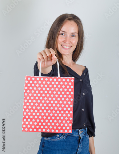Young woman holding paper bag in black shirt, jeans shorts and looking cheerful. front view.