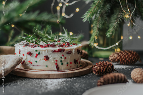 Christmas cake with white cheese cream, decorated with cranberries and rosemary. New Year's treat on the background of fir branches and garlands.