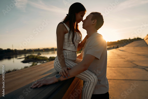 A young couple is sitting on a bench at sunset. The lady sits on the guy's lap and looks so contented and happy.