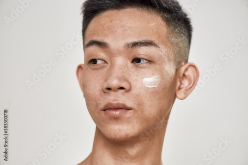 Close up portrait of young asian man with problematic skin applied cream on his face, looking aside isolated over white background. Beauty, skincare, treatment concept photo