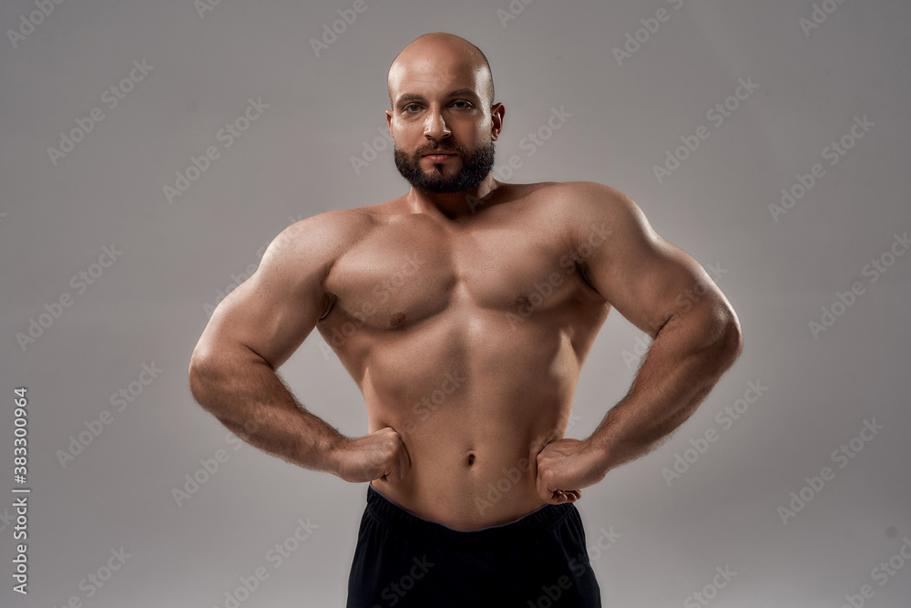 Your personal fitness trainer. Muscular strong caucasian man bodybuilder showing his perfect body looking at camera while posing shirtless isolated over grey background