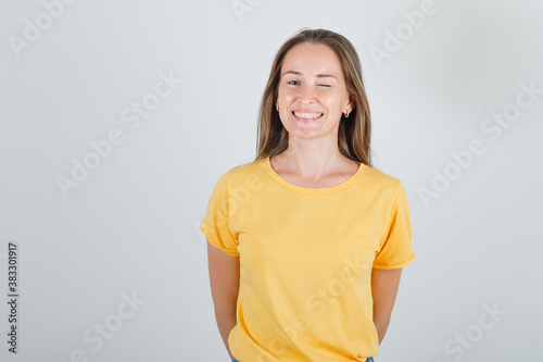 Young woman in yellow t-shirt winking and holding hands behind her and looking merry , front view.