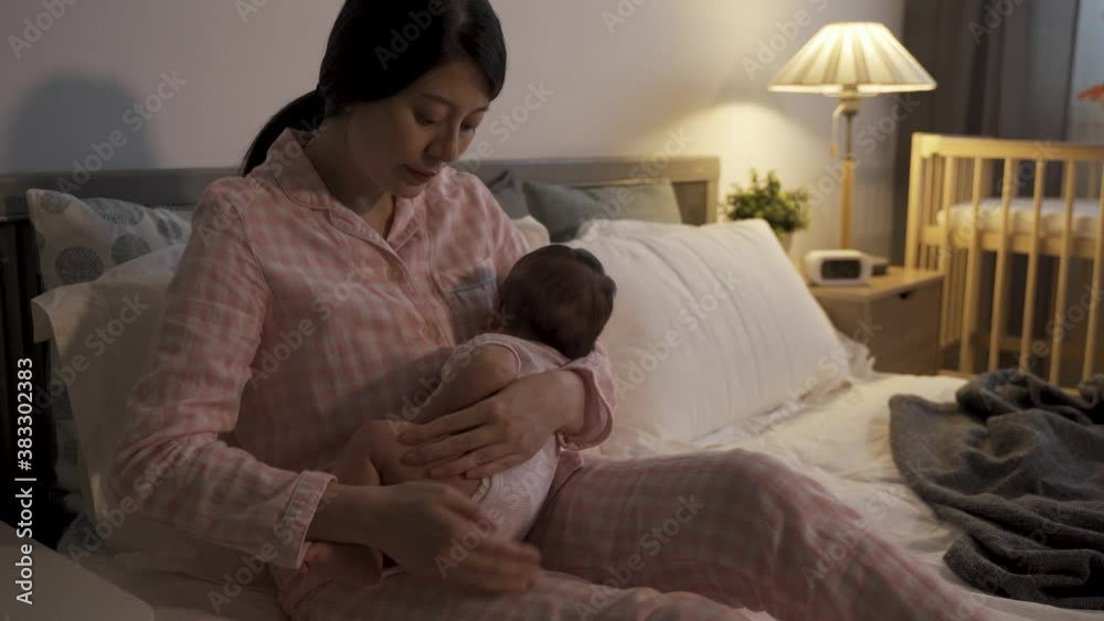 cropped shot of asian new mother sitting on bed is patting and coaxing her wailing baby at night during bedtime in the bedroom at home. cropped shot of asian new mother sitting on bed is patting and coaxing her wailing baby at night during bedtime in the bedroom at home.