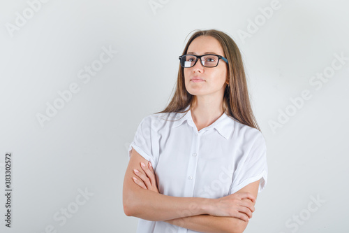 Young woman looking away with crossed arms in white t-shirt, glasses front view.