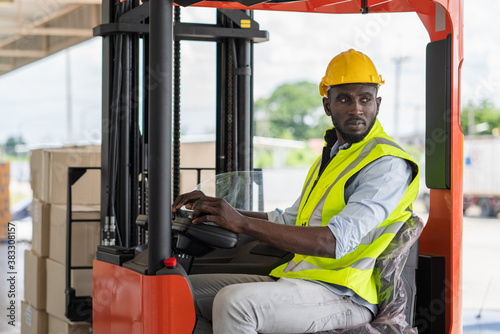 Male worker in safety vest and yellow helmet driving a forklift at warehouse factory