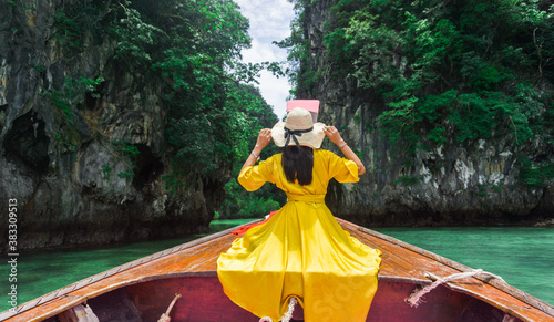 Woman wearing a long yellow dress, holding her hands on her hat, sitting back on a local long-tail boat, she travels to Hong Island, a beautiful sea in Krabi, Thailand.