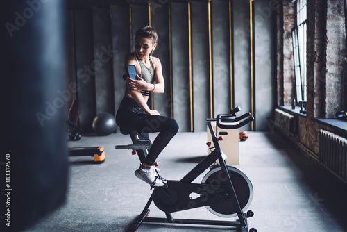 Young caucasian sports women in activewear sitting on exercising bike looking at smartphone choosing playlist songs, pretty female athlete looking at mobile phone on arms during workout