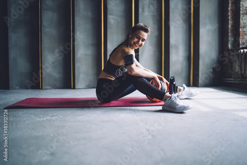 Happy pretty girl in activewear with smartphone holder on arms listening music during workout, smiling caucasian sportswoman using mobile phone enjoying playlist songs while training with ball