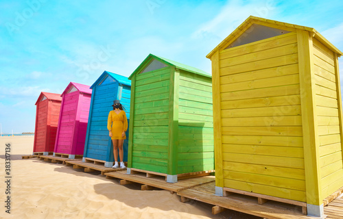 Girl with mask and glasses on the beach with the colored huts next to it. New normal concept.