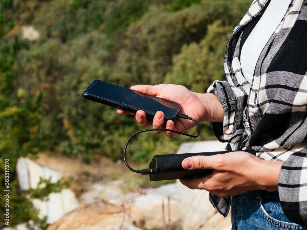 Fototapeta premium The black smartphone is connected to the power Bank. Portable chargers and external batteries in everyday life. A girl holds a phone and a powerBank against the background of nature. Tourism concept
