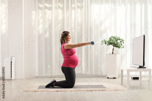 Pregnant woman exercising with blue dumbbells in front of a tv