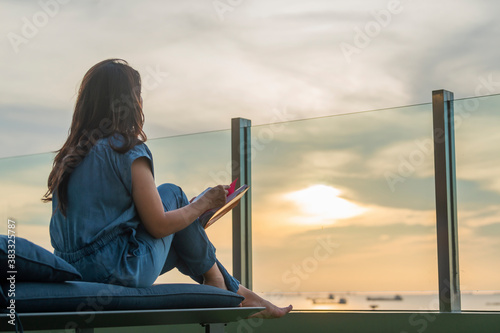 A portrait of beautiful asian woman holding book in hand and enjoying peaceful moment of beautiful colorful sunset.