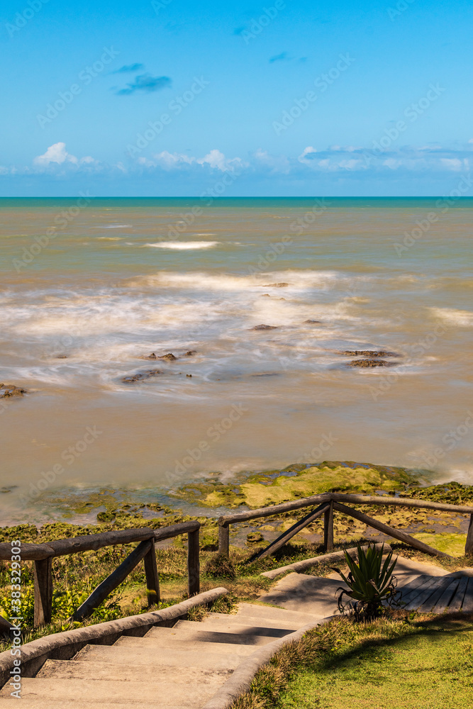 View from the top of a wooden and stone staircase of the blurred sea, the reef and a blue sky.