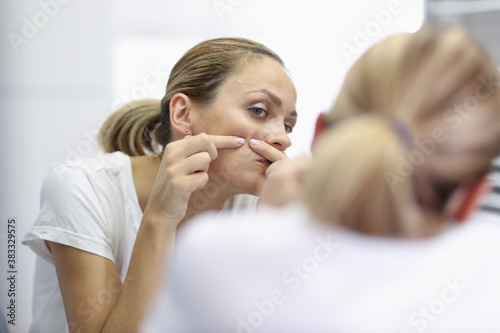 Young woman stands in front of mirror and presses pimples with hands portrait. Facial skin care cleaning at home concept. © H_Ko
