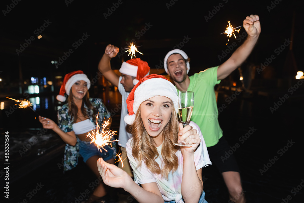 Selective focus of woman in santa hat holding glass of champagne and sparkler near friends outdoors at night Selective focus of woman in santa hat holding glass of champagne and sparkler near friends outdoors at night