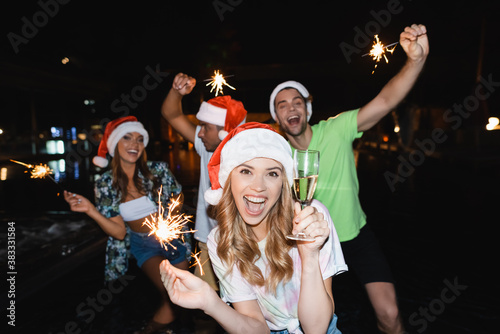 Selective focus of woman in santa hat holding glass of champagne and sparkler near friends outdoors at night
