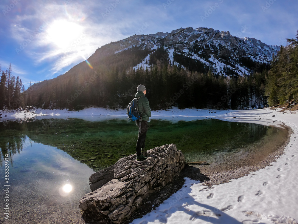 A man standing on a big stone by the shore of Green Lake in Austrian Alps in winter. Snow covers the ground. Crystal clear water. Reflection of Alps in calm lake's water. Winter wonderland. Adventure