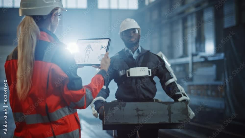 Female Engineer is Testing Vital Components of a Futuristic Bionic Exoskeleton that Her Project Assistant is Wearing in a Metal Industry Factory. Contractor is Lifting Steel Parts in a Powered Shell. Female Engineer is Testing Vital Components of a Futuristic Bionic Exoskeleton that Her Project Assistant is Wearing in a Metal Industry Factory. Contractor is Lifting Steel Parts in a Powered Shell.