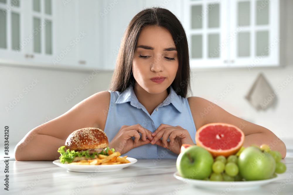 Woman choosing between fruits and burger with French fries in kitchen Woman choosing between fruits and burger with French fries in kitchen