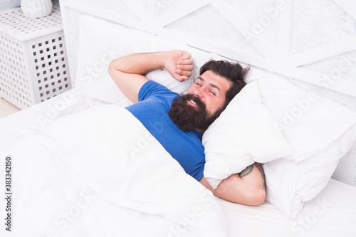 Photo of handsome cheerful woke up man lies in bed indoors at home looking camera, relax and recreation