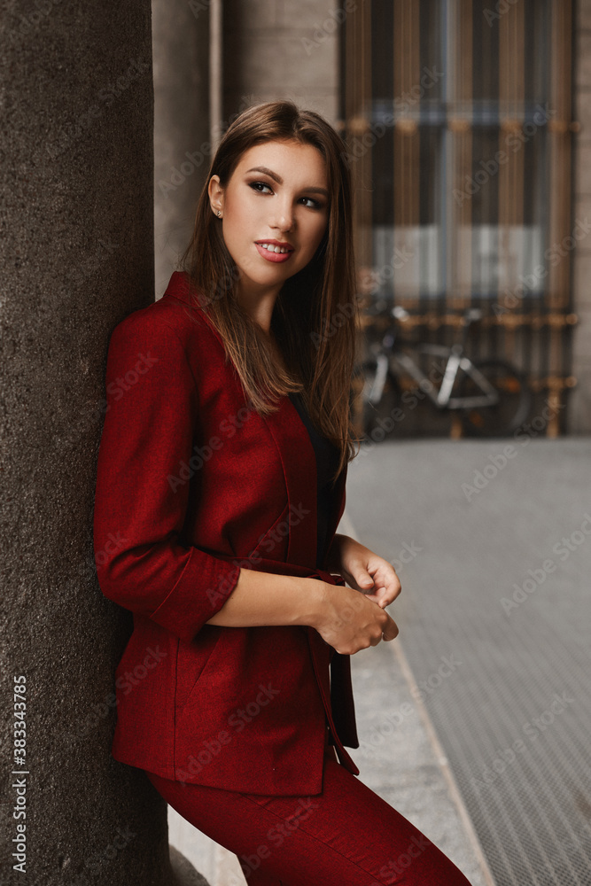 A young woman with a broad smile in a red fashionable suit looking aside and posing outdoors in the city street A young woman with a broad smile in a red fashionable suit looking aside and posing outdoors in the city street