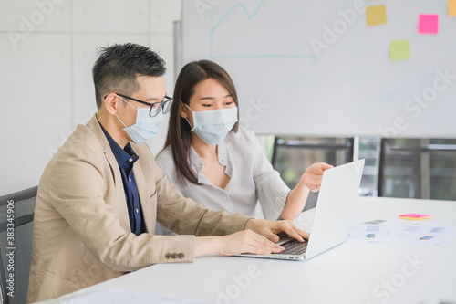 Asian businessman and businesswoman wears face mask for protect coronavirus discuss business project in laptop in meeting room at modern office. image focused on businessman