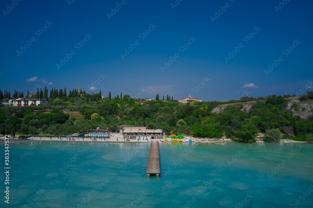 Island against the blue sky. Wooden pier, turquoise water. Panoramic aerial view of Lido delle Bionde beach, Sirmione, Lake Garda, Italy.