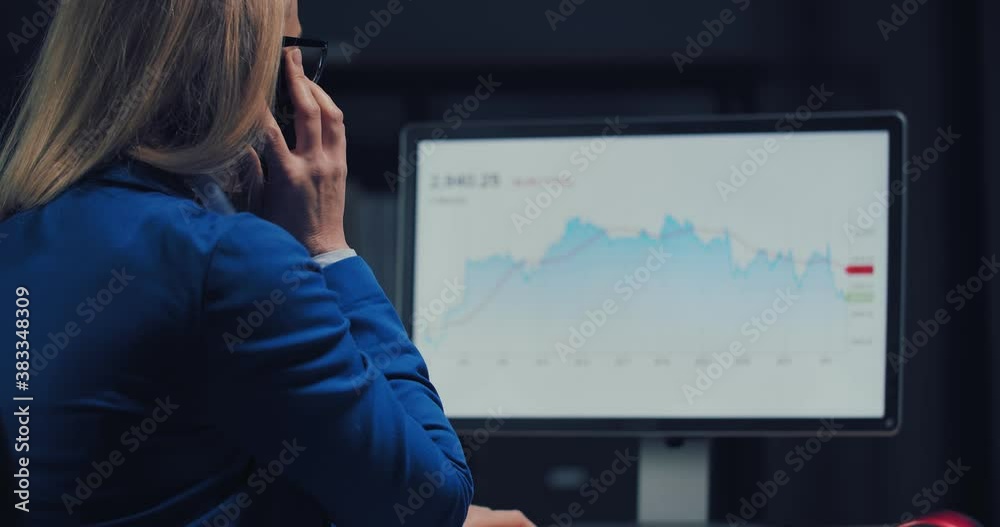 View from shoulder of business woman talking on smartphone while analyzing financial statistic on computer. Dark atmosphere at modern office. View from shoulder of business woman talking on smartphone while analyzing financial statistic on computer. Dark atmosphere at modern office.