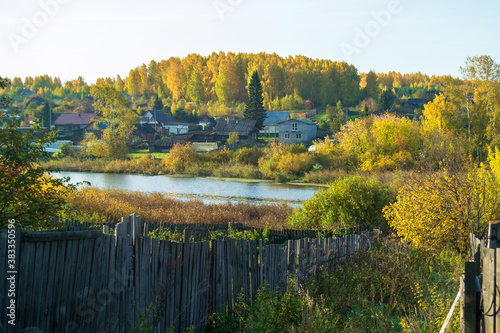 Beautiful autumn landscape. Village on the river Bank with colorful trees, against the blue sky. Old wooden fence in the foreground.