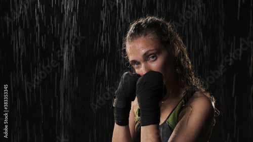 Nice athletic woman in boxing bandages and green and gray sports top fights with camera. A sports lady in the rain at night wrestles and hits the camera. Rack focus.