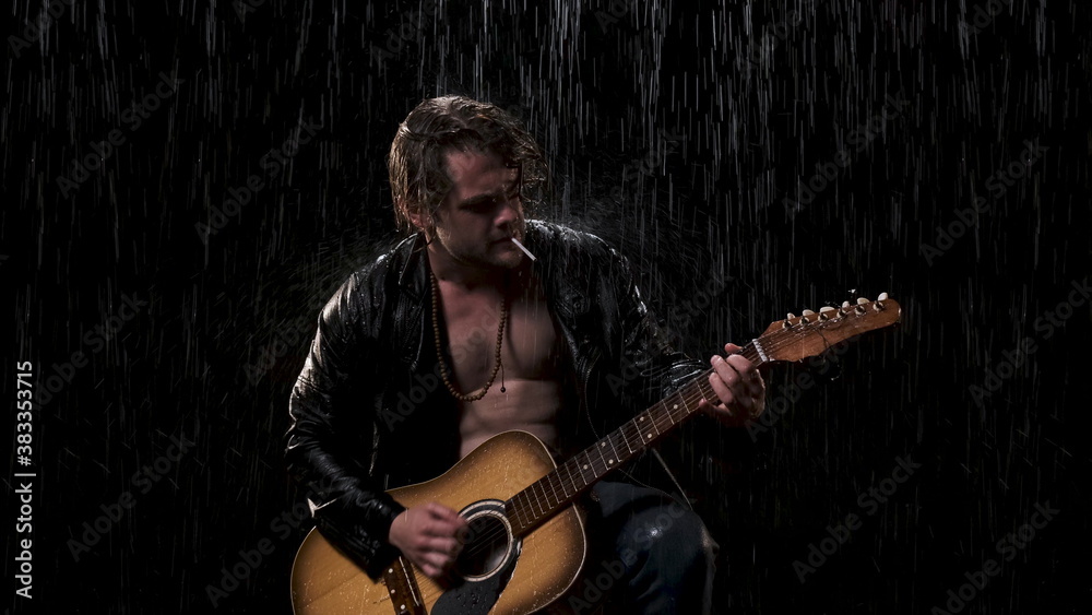 Obraz premium long hair sexy guitarist in black leather jacket plays acoustic guitar and sings song on black background in the rain outside at night.